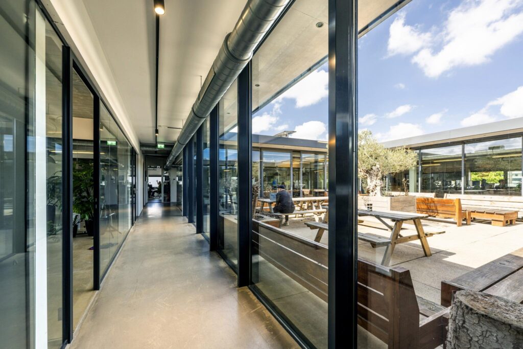 Modern office hallway with glass walls overlooking a sunny courtyard with wooden picnic tables on Johan van Hasseltweg.